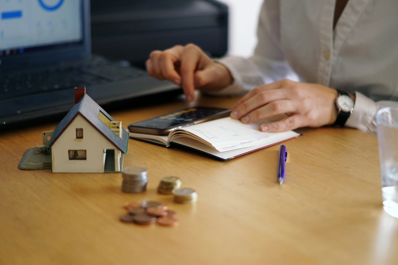 Person calculating home budget with smartphone calculator beside model house and stacked coins on desk