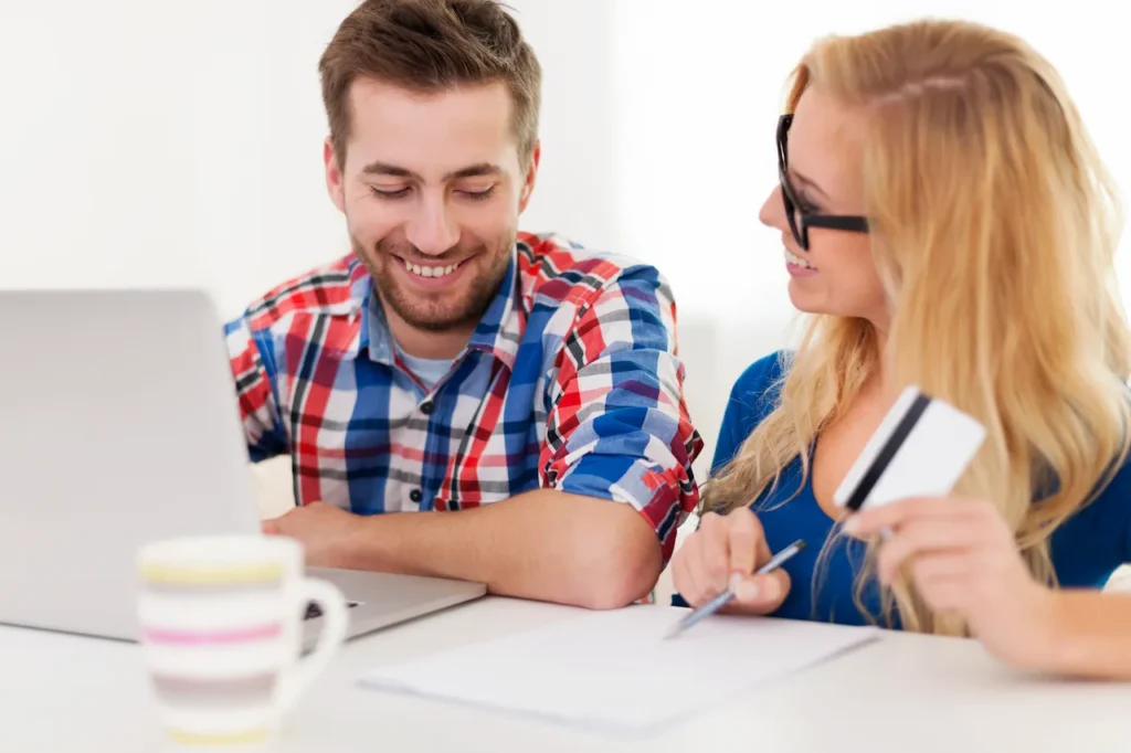 Couple sitting at a table using a laptop to pay bills online, woman holding a credit card and pen while reviewing paperwork.