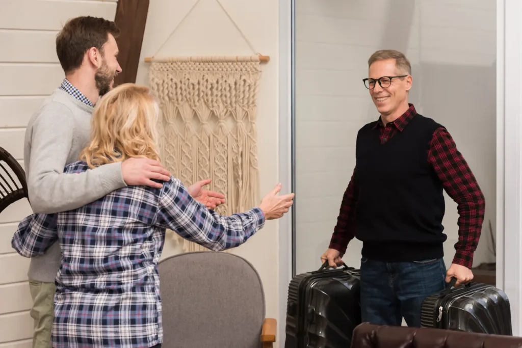 Smiling man arriving with suitcases while a couple greets him warmly inside a home, preparing to welcome a guest.