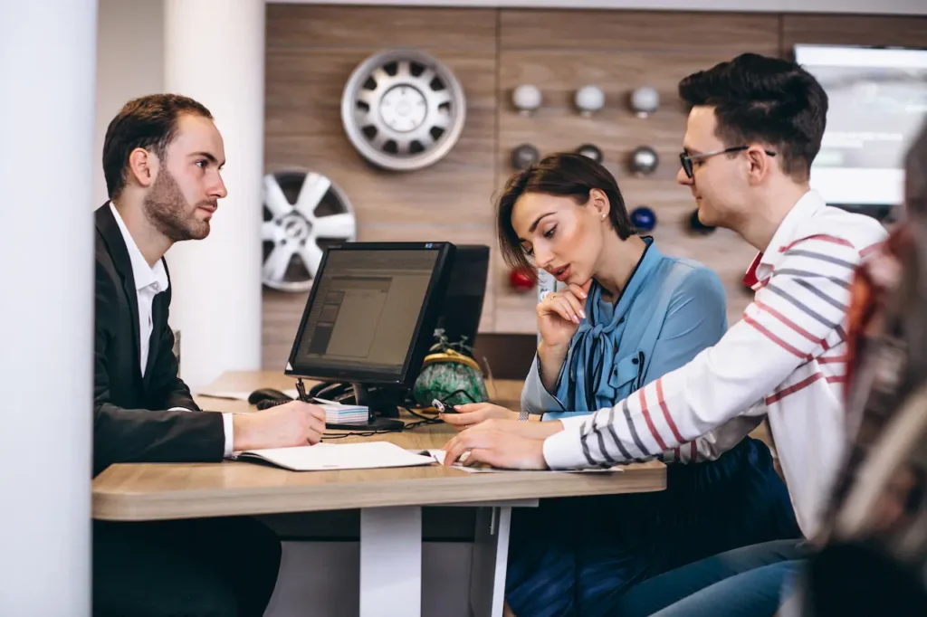 Car dealer discussing paperwork with a couple at a desk in a showroom, with vehicle wheel displays visible in the background.