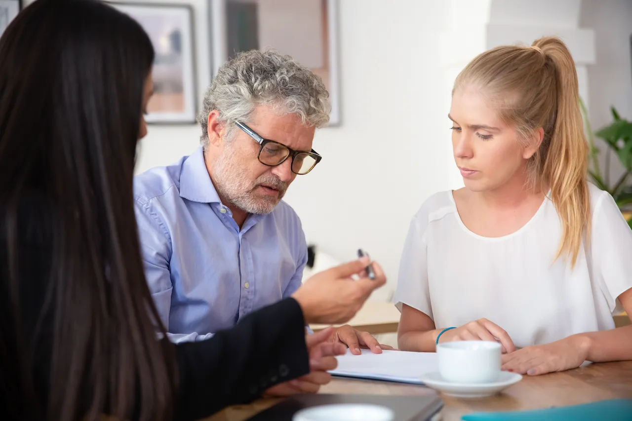 Business meeting scene with a mature man reviewing documents while two women sit beside him, one handing him a pen to sign paperwork.