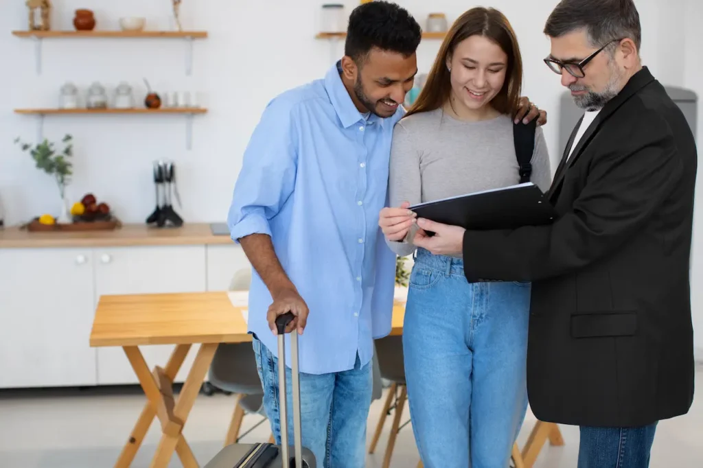 Couple standing with a travel suitcase while reviewing documents with a man in a suit inside a modern home setting.