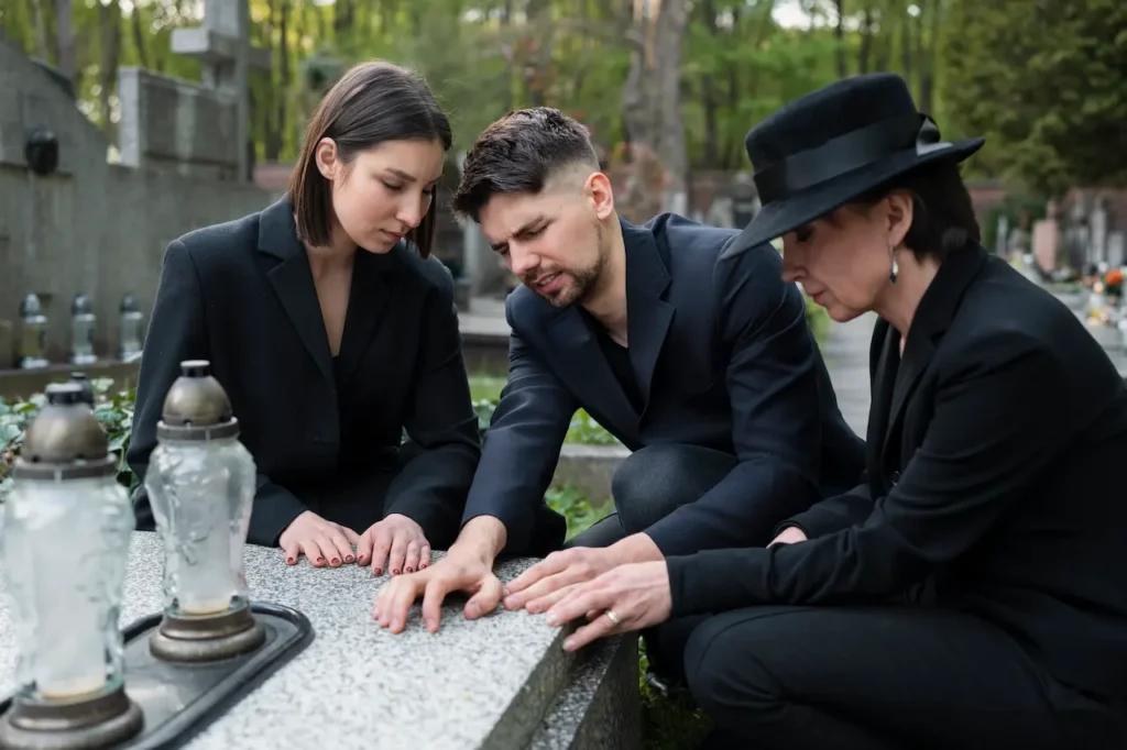 Three people dressed in black kneeling together and touching a gravestone at a cemetery