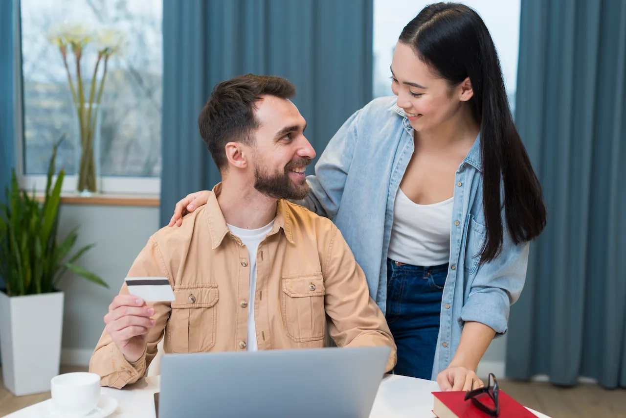 Smiling couple shopping online at home, man holding a credit card while using a laptop, woman standing beside him and looking at the screen.