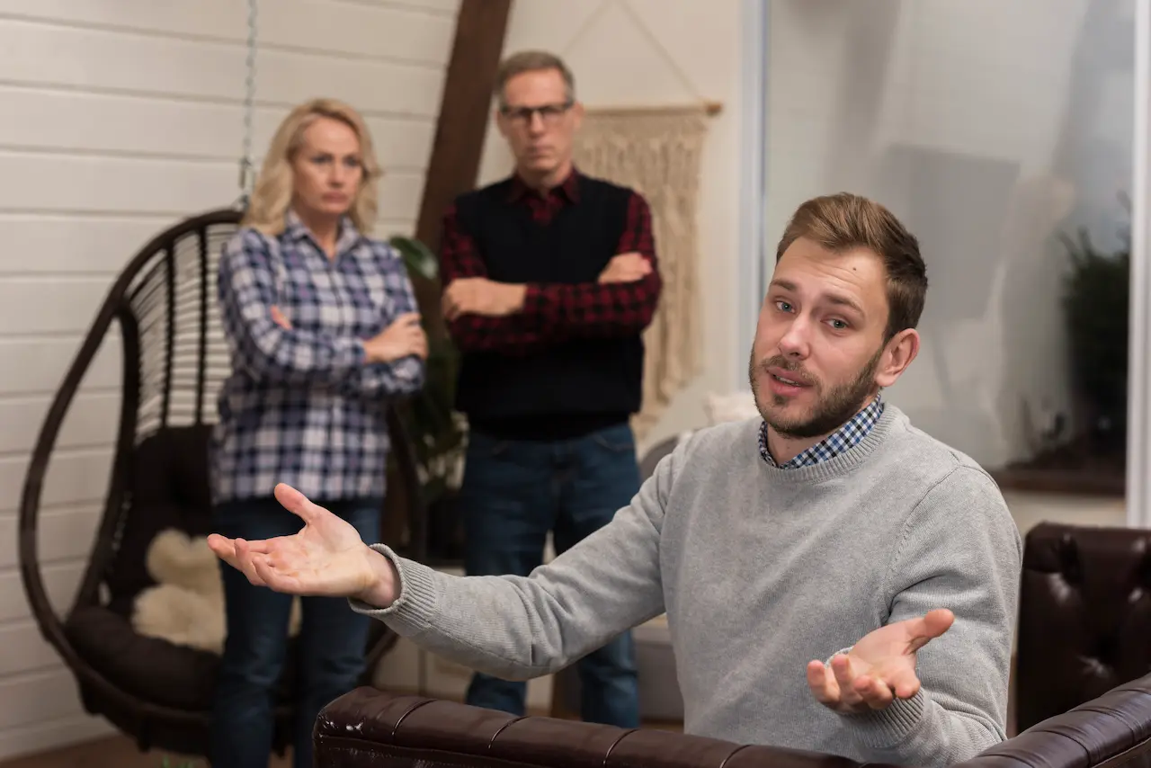 Young man sitting on a sofa gesturing with his hands while two older adults stand behind him with arms crossed, appearing upset during a tense discussion at home.