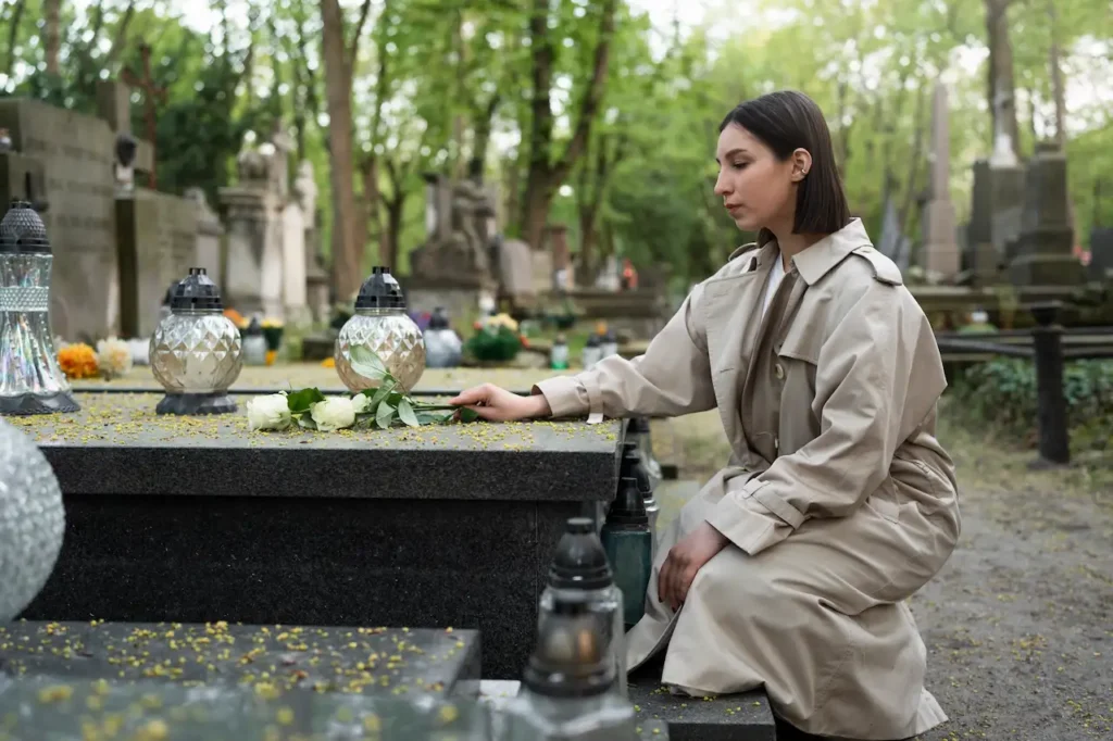 Woman placing white roses on a grave in a cemetery, kneeling beside a headstone surrounded by memorial candles.
